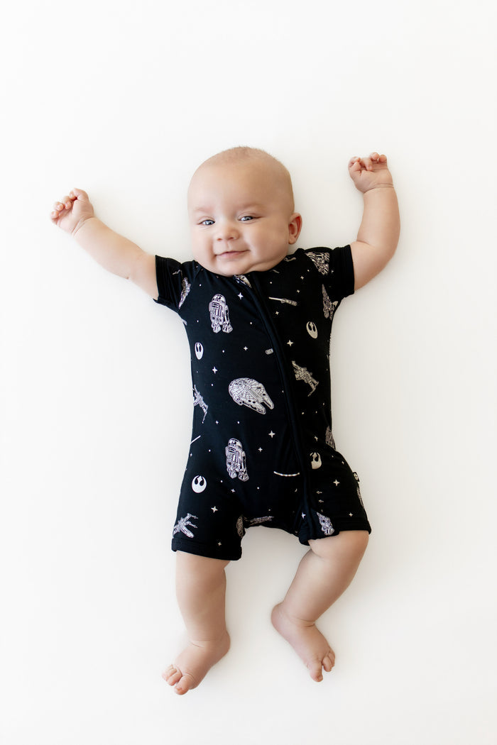 Infant stretched out on a white surface wearing the Zipper Shortall Romper in STAR WARS™
