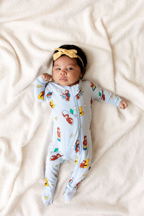 Baby wearing Blue Zippered Footie in Curious George print lying on beige sheet