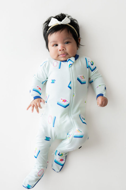 Baby wearing Zippered Footie in Dew Mahjong print and a bow headband on a beige background