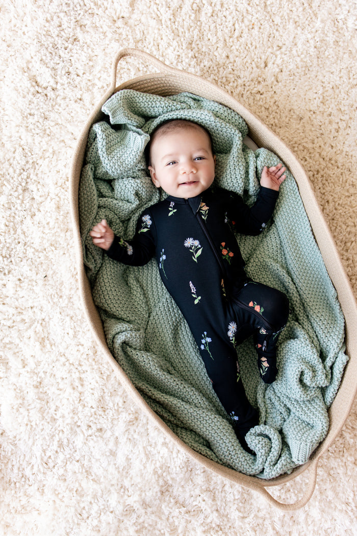 Baby lying on green blanket in a basket wearing Zippered Footie in Midnight Wildflower print