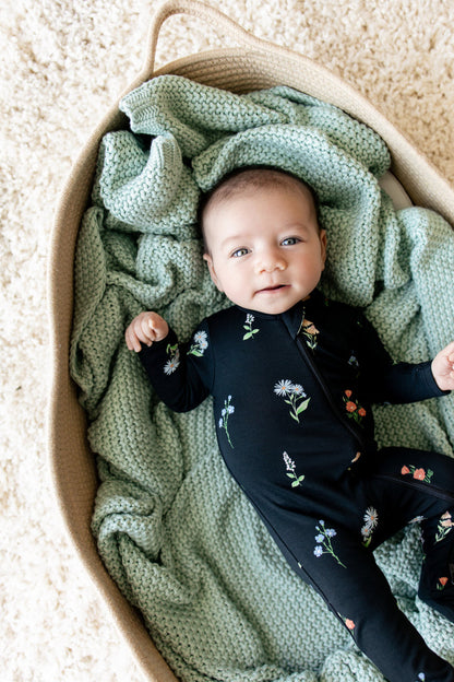 Baby lying on green blanket in a basket wearing Zippered Footie in Midnight Wildflower print